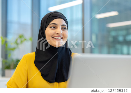 A smiling woman wearing a black hijab looks towards the right in a modern office setting. She is wearing a yellow shirt. 127459355