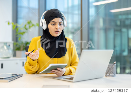 A woman wearing a hijab participates in a video conference, engaged in a discussion while seated at her desk. A woman wearing a hijab participates in a video conference, engaged in a discussion while seated at her desk. 127459357