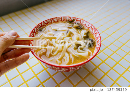person is eating noodles with chopsticks. colorful bright red plate of ramen sits on yellow table. person is eating noodles with chopsticks. colorful bright red plate of ramen sits on yellow table. 127459359