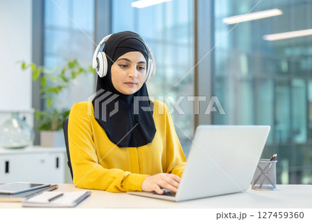 An observant Muslim woman wearing a hijab and headphones is working on a laptop in a modern office setting, possibly attending an online class. 127459360