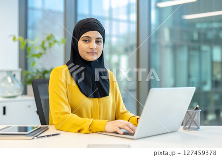 A woman wearing a hijab sits at a desk working on a laptop in a modern office setting. She is focused and appears to be working professionally. 127459378