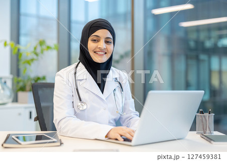 A smiling female doctor wearing a hijab sits at a desk, working on a laptop in a modern office setting, focusing on her work with a friendly demeanor. 127459381