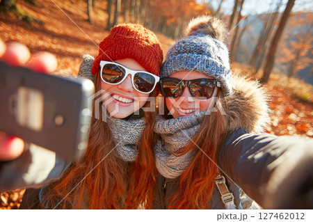 Two young women taking a selfie in a vibrant autumn forest with colorful leaves surrounding them 127462012