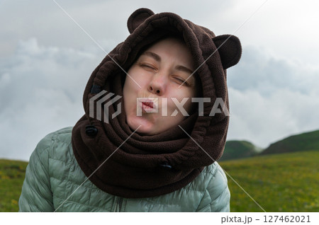 Cute young woman traveler in down sweater and fleece hoodie with bear ears blowing kiss to camera with closed eyes against natural evening landscape Cute young woman traveler in down sweater and fleece hoodie with bear ears blowing kiss to camera with closed eyes against natural evening landscape 127462021