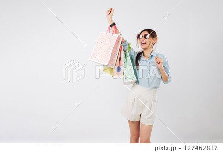 Happy young Asian woman shopping on Black Friday raising her hand with shopping bags, cheerful and excited holding colorful bags after shopping, isolated studio white background 127462817
