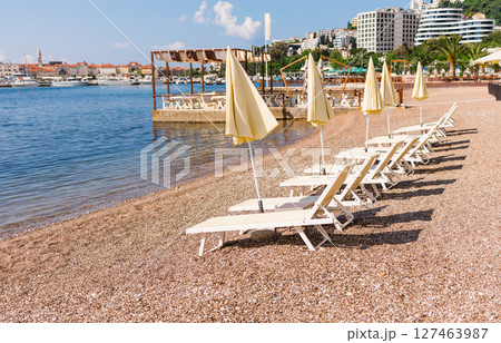 Empty sun loungers and closed umbrellas on the Adriatic coast. Summer beach infrastructure and seaside tourism in Budva, Montenegro Empty sun loungers and closed umbrellas on the Adriatic coast. Summer beach infrastructure and seaside tourism in Budva, Montenegro 127463987