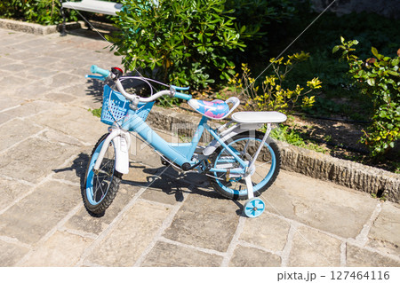 Blue kids bicycle with training wheels on sunny pavement. Childhood fun, outdoor learning, and the joy of early cycling experiences in a quiet garden path. Blue kids bicycle with training wheels on sunny pavement. Childhood fun, outdoor learning, and the joy of early cycling experiences in a quiet garden path. 127464116