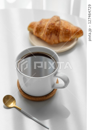 Cup of espresso coffee and fresh croissant on white table near window with morning shadows. Cup of espresso coffee and fresh croissant on white table near window with morning shadows. 127464729