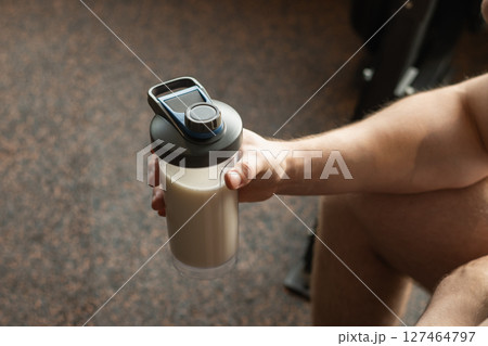 Close up shot of bottle with holding protein shaker in mans hand at gym. Bodybuilder during strength training. Fitness healthy lifestyle concept. Supplement product, creatine, glutamine for workout. 127464797