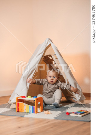 Cheerful 2 year old boy sitting near the wigwam and playing with a wooden toy. Toddler boy in a beige t-shirt sitting in the living room and playing with toys. Child in comfortable home clothes 127465700