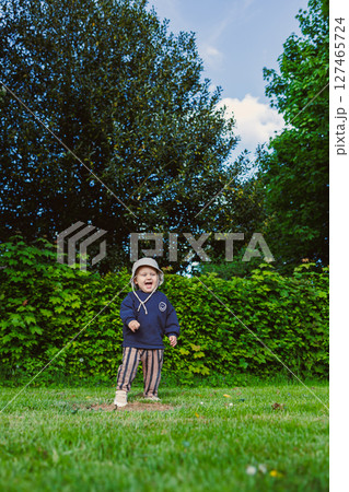 Stylish smiling toddler boy in a panama hat on his head runs on green grass in a city park. A walk of a 2 year old boy in nature. The child enjoys the beauty of nature and a sunny day in summer. 127465724