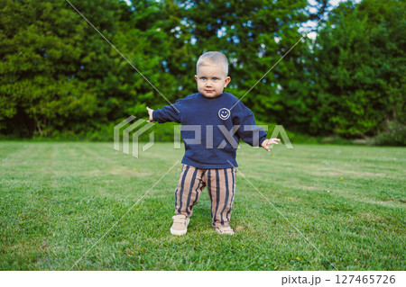 Toddler laughing and enjoying warm sunny day. Happy 2 year old boy in t-shirt and shorts running on green grass and laughing outdoors. Child playing on public playground on sunny summer day. 127465726
