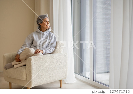 Peaceful mature woman in hoodie sitting in cozy chair looking out large window at home interior Peaceful mature woman in hoodie sitting in cozy chair looking out large window at home interior 127471716