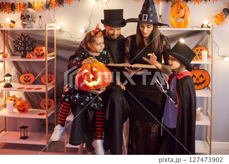 Family With Children Celebrating Halloween Party At Home Reading A Book Together In Costumes 127473902