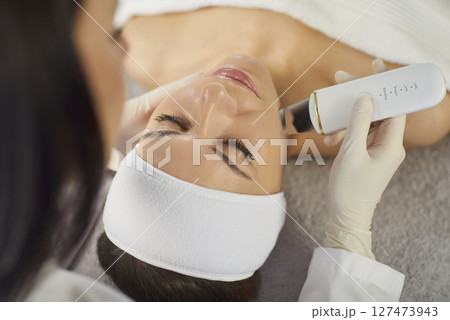 Beautician performing ultrasonic facial cleansing with ultrasonic device on woman during treatment. Beautician performing ultrasonic facial cleansing with ultrasonic device on woman during treatment. 127473943