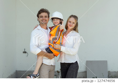 A family smile while standing on a construction site. The father hold their child in a safety vest and helmet while the mother stand beside them, both wearing casual clothing. A family smile while standing on a construction site. The father hold their child in a safety vest and helmet while the mother stand beside them, both wearing casual clothing. 127474056