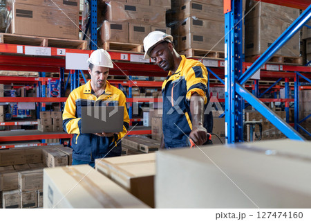 Two warehouse worker in safety uniform review inventory record using barcode scanner and laptop. Stand among stacked box in a busy storage area. Working atmosphere in warehouse with racking system. Two warehouse worker in safety uniform review inventory record using barcode scanner and laptop. Stand among stacked box in a busy storage area. Working atmosphere in warehouse with racking system. 127474160