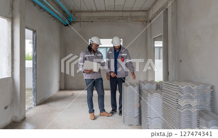 Two construction worker in safety gear are inspecting roofing material in an incomplete building. Natural light illuminate the spacious area filled with stack of tile. 127474341