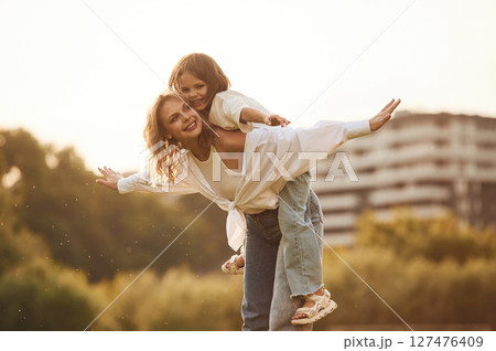 Sitting on the mother shoulders. Woman with her little daughter are on the summer field together Sitting on the mother shoulders. Woman with her little daughter are on the summer field together 127476409