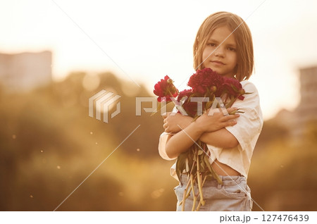 Holding beautiful red flowers. Little girl is on the summer field at daytime 127476439