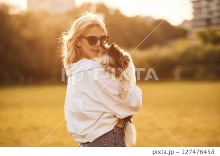 Woman in sunglasses is standing on the summer field and holding little dog 127476458