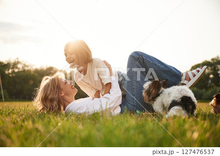 Laying down on ground. Beautiful mother with her little daughter and dogs are on the summer field together 127476537