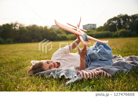 Laying down on the ground. Happy little girl is playing with toy plane outdoors Laying down on the ground. Happy little girl is playing with toy plane outdoors 127476608