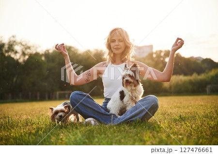 Sitting in yoga lotus pose. Young woman is with two little dogs on the summer field Sitting in yoga lotus pose. Young woman is with two little dogs on the summer field 127476661