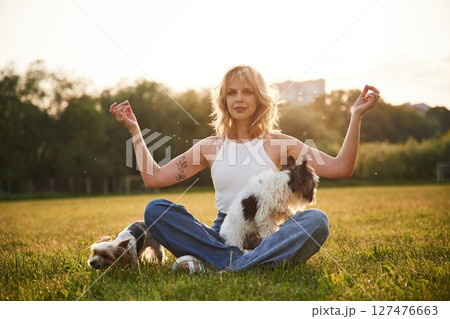 Sitting in yoga lotus pose. Young woman is with two little dogs on the summer field 127476663