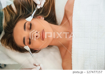 Top view, using brushes. Woman face getting facial care by beautician hands at spa salon 127477157