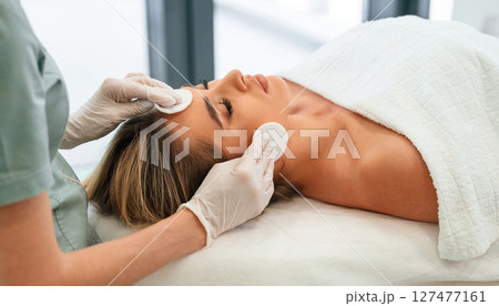 Cleaning skin by using cotton pad or sponge. Woman face getting facial care by beautician hands at spa salon 127477161