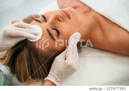 Cleaning skin by using cotton pad or sponge. Woman face getting facial care by beautician hands at spa salon Cleaning skin by using cotton pad or sponge. Woman face getting facial care by beautician hands at spa salon 127477163