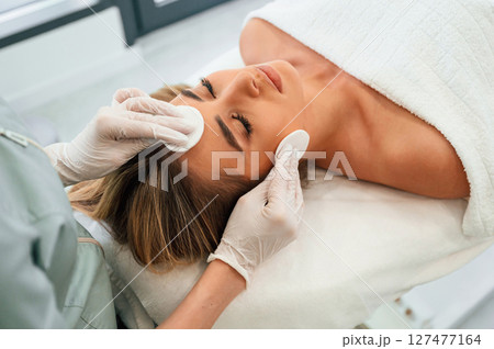 Cotton pad or sponge are used to clean the skin. Woman face getting facial care by beautician hands at spa salon 127477164