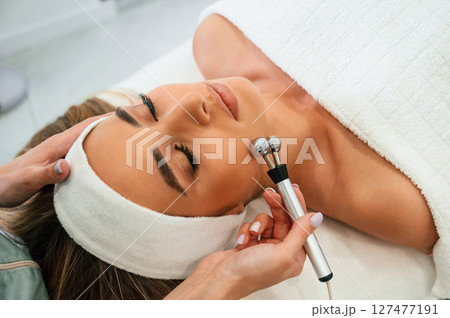 Massage roller is used. Woman face getting facial care by beautician hands at spa salon Massage roller is used. Woman face getting facial care by beautician hands at spa salon 127477191