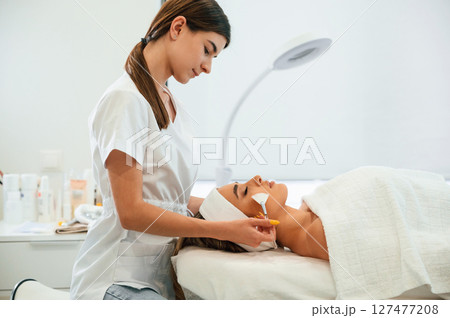 Lying down. Using brushes. Woman face getting facial care by beautician hands at spa salon 127477208