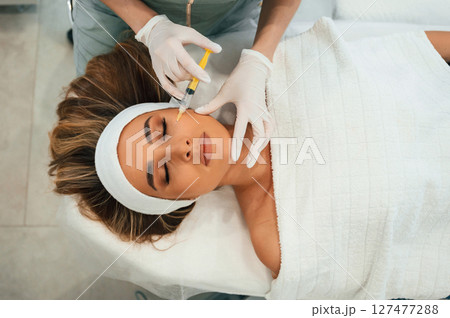 Yellow colored syringe, making injection. Woman face getting facial care by beautician hands at spa salon 127477288