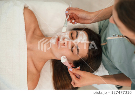 Applying clay face mask. Woman face getting facial care by beautician hands at spa salon Applying clay face mask. Woman face getting facial care by beautician hands at spa salon 127477310