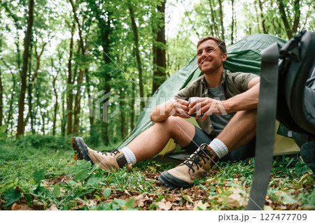 With cup of water. Sitting near the tent. Tourist in summer forest. Conception of exploration and leisure 127477709