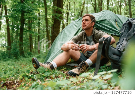 With cup of water. Sitting near the tent. Tourist in summer forest. Conception of exploration and leisure 127477710
