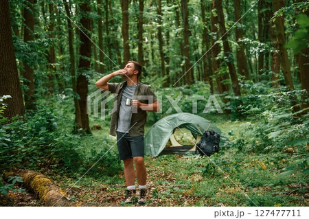 Green colored tent is behind. Tourist in summer forest. Conception of exploration and leisure Green colored tent is behind. Tourist in summer forest. Conception of exploration and leisure 127477711