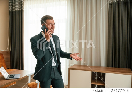 Talking by phone. Businessman is indoors in the hotel room Talking by phone. Businessman is indoors in the hotel room 127477768
