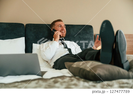 Having fun by talking by phone and lying down on bed. Businessman is indoors in the hotel room Having fun by talking by phone and lying down on bed. Businessman is indoors in the hotel room 127477789