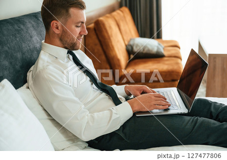 Side view. Lying down on the bed with laptop. Businessman is indoors in the hotel room 127477806