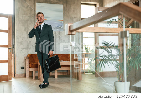 Beautiful man in suit and tie is standing indoors and holding case in hand 127477835