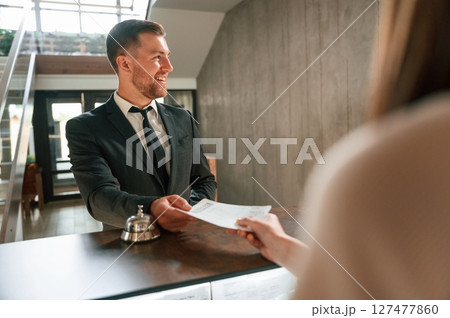 Woman serves a visitor. A man in a black jacket at the hotel reception Woman serves a visitor. A man in a black jacket at the hotel reception 127477860