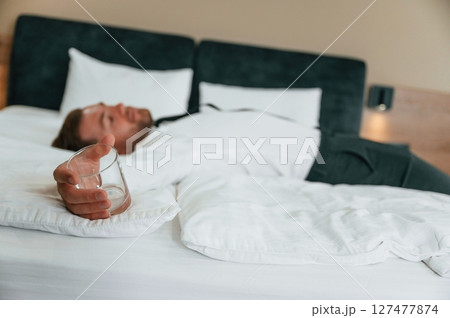 Empty glass in hand, lying down on the bed, tired. Businessman is indoors in the hotel room 127477874