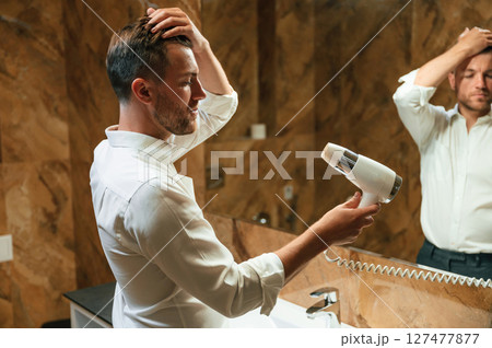 Drying his hair. Man in formal clothes is in the bathroom against mirror 127477877