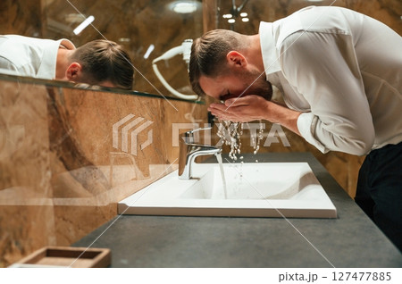 Washing the face. Man in formal clothes is in the bathroom against mirror 127477885