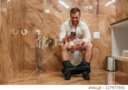 Watching funny content. A man in a white shirt is sitting on the toilet, holding a smartphone in his hands 127477892
