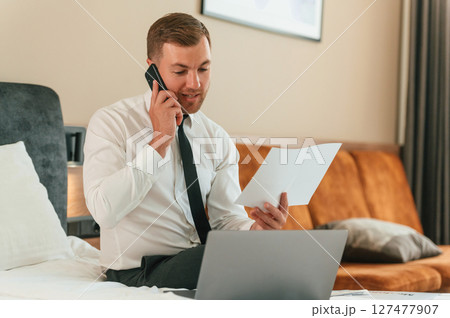 Reading the information from ticket, talking by phone. Businessman is indoors in the hotel room Reading the information from ticket, talking by phone. Businessman is indoors in the hotel room 127477907